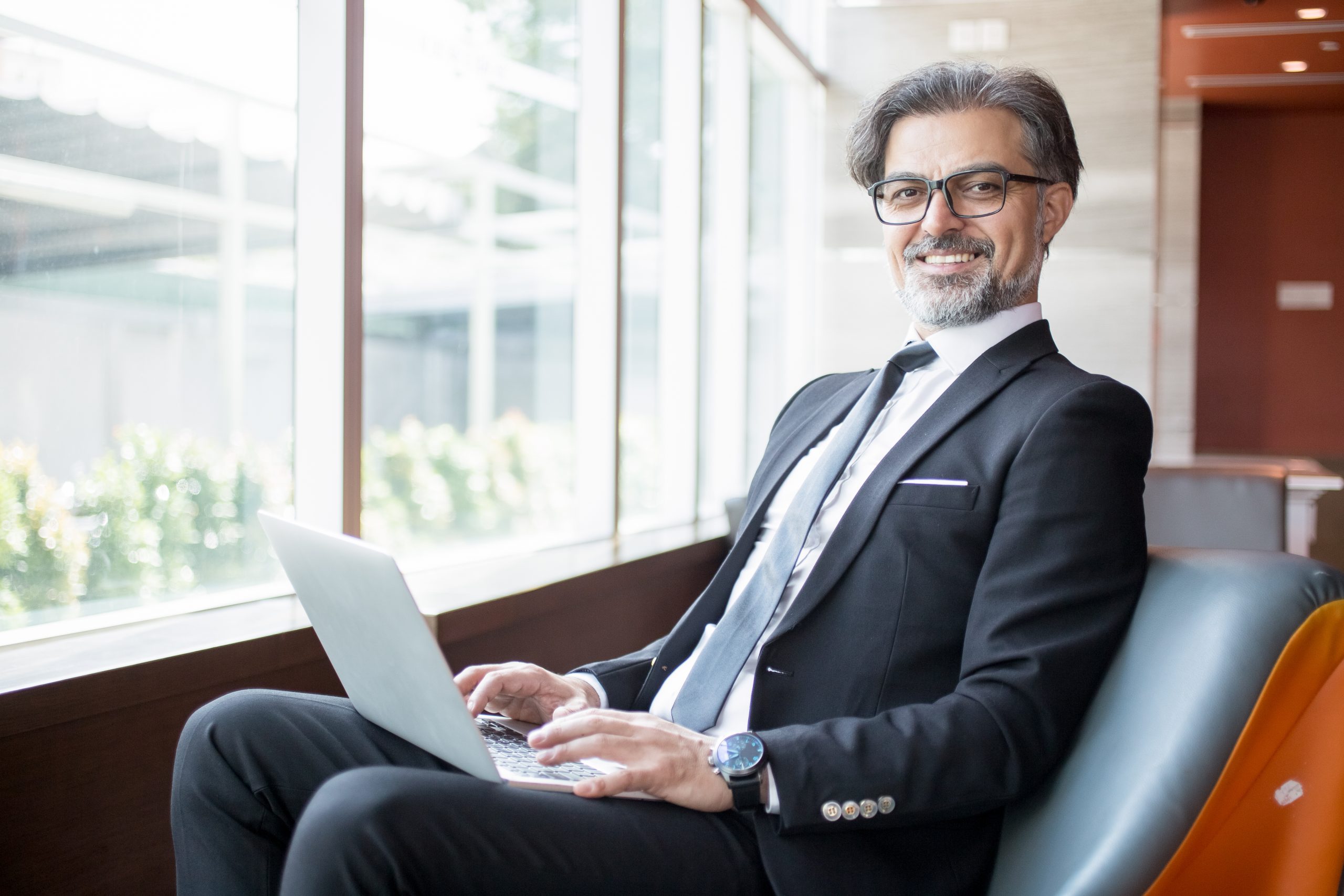Closeup portrait of smiling middle-aged handsome business man looking at camera and working on tablet computer on his lap at window in lobby
